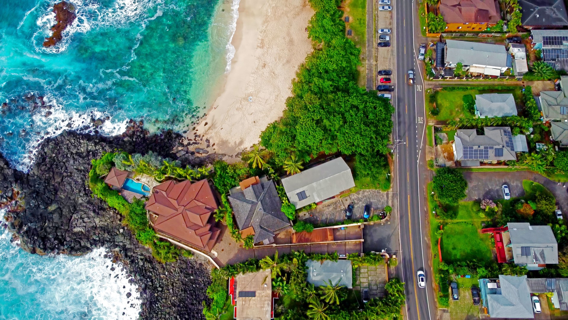 Aerial View of Luxury Beachfront Homes in Hawaii. A breathtaking aerial shot of beachfront houses along a tropical coastline in Hawaii, featuring turquoise ocean waves, sandy beach, and lush greenery.